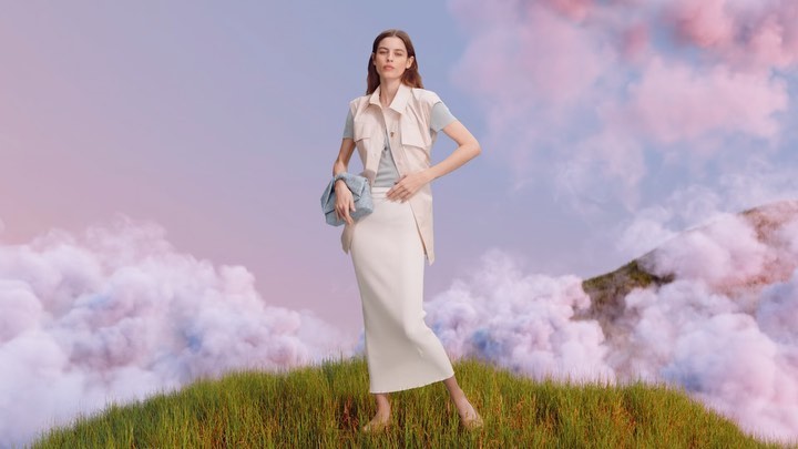 Fashion shoot of a young adult woman wearing a skirt and vest, with a blue purse, standing on a grassy hill against a cloudscape background.