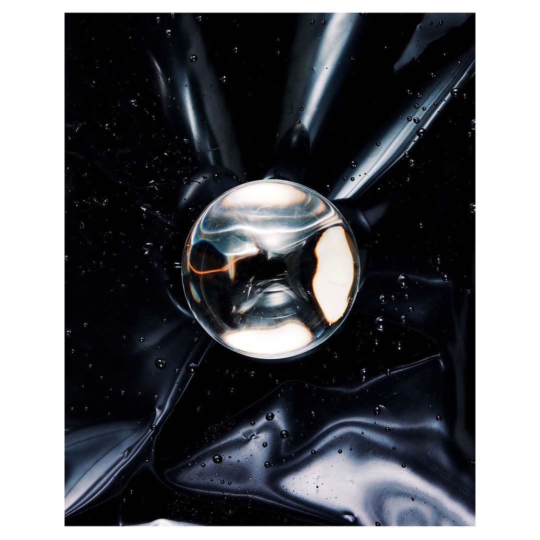 Abstract still life of a clear glass orb resting on shiny black plastic, reflecting light with subtle water droplets on the surface.