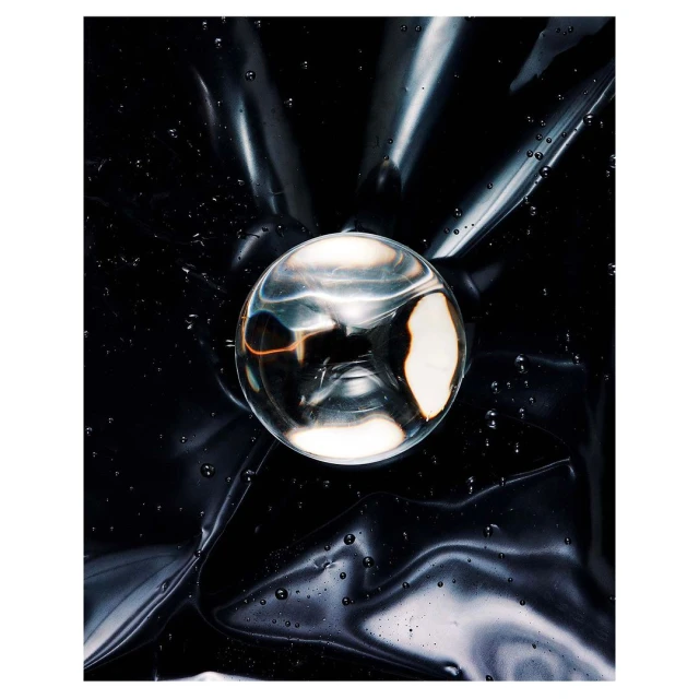 Abstract still life of a clear glass orb resting on shiny black plastic, reflecting light with subtle water droplets on the surface.