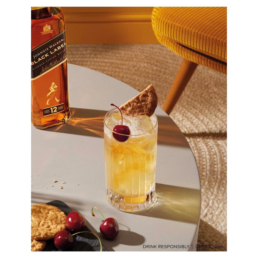 Whiskey cocktail with a cherry garnish and a bottle of Johnnie Walker Black Label, sitting on a grey table next to a mustard chair.