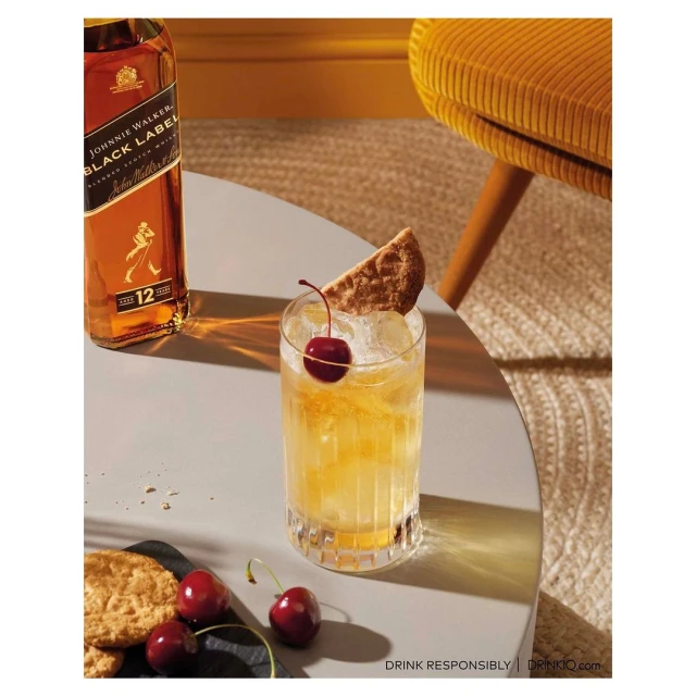 Whiskey cocktail with a cherry garnish and a bottle of Johnnie Walker Black Label, sitting on a grey table next to a mustard chair.