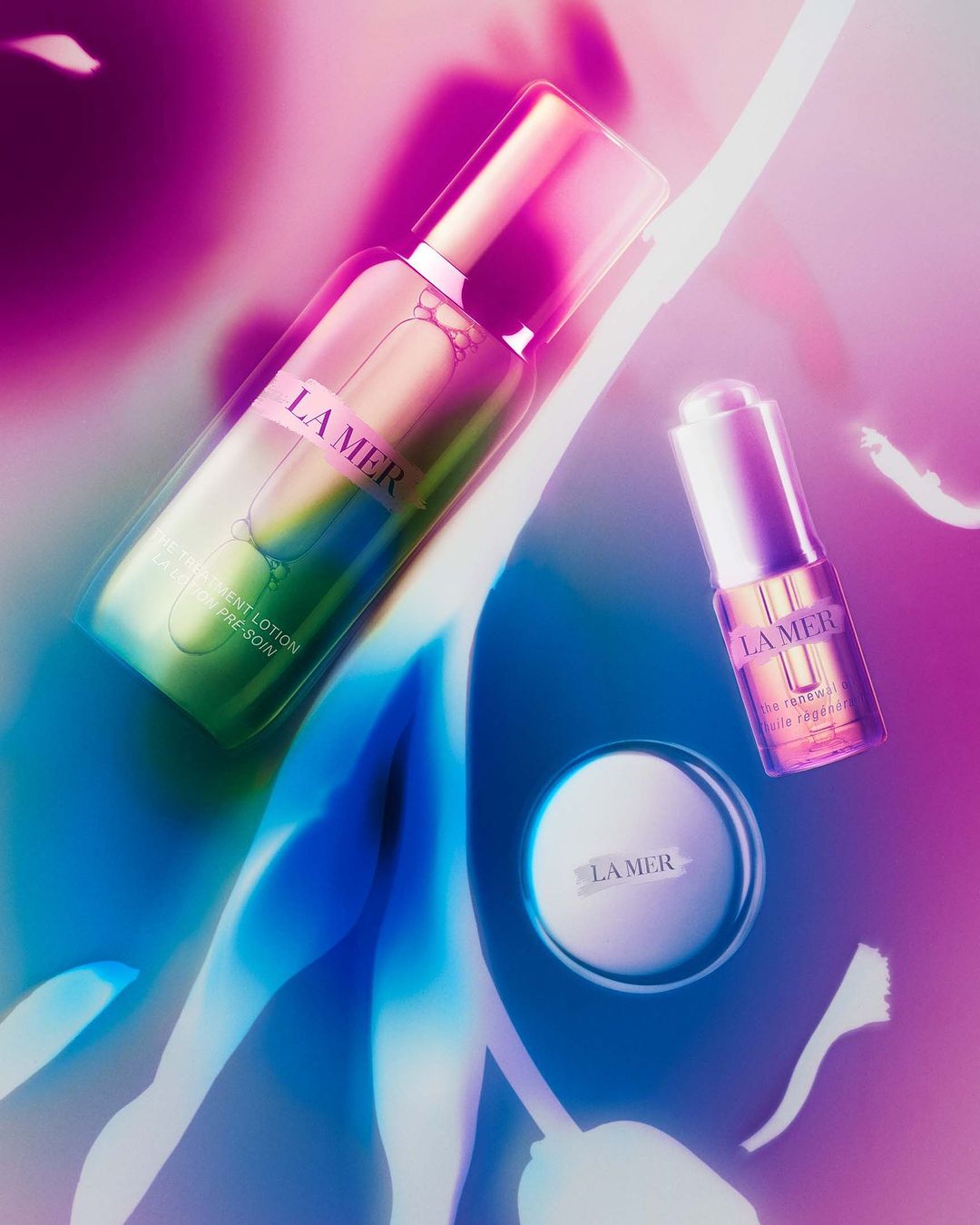 Three La Mer skincare products displayed on a multicolored surface, including lotion, cream, and serum with pink and blue lighting.