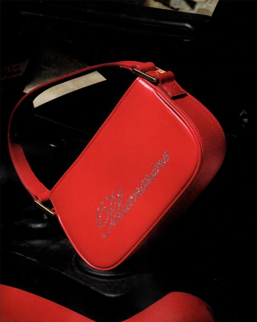 Close-up studio shot of a small red leather purse with a shoulder strap and a crystal logo, resting on a shiny black surface.
