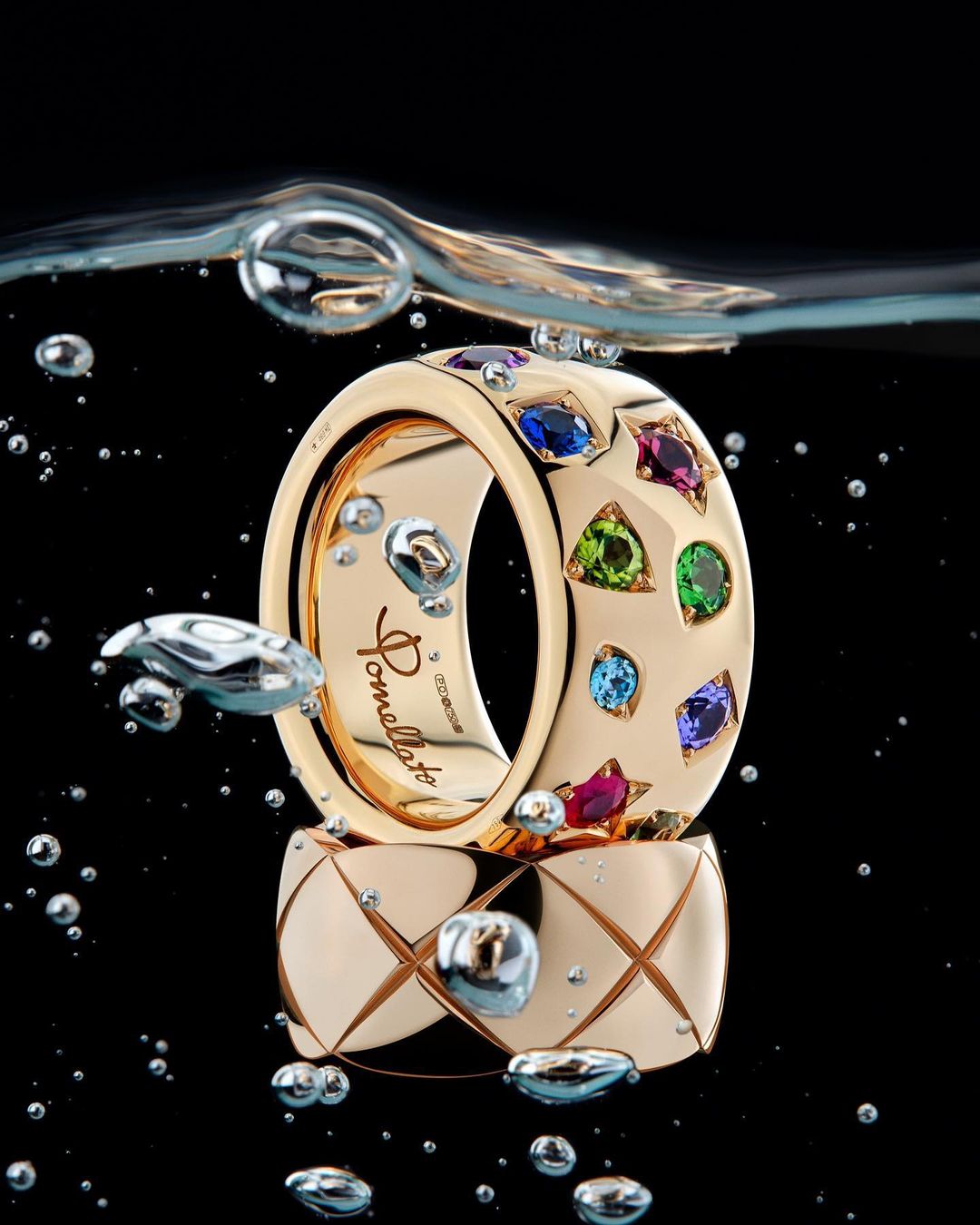 Two gold rings set with colorful gemstones submerged underwater with air bubbles against a black background