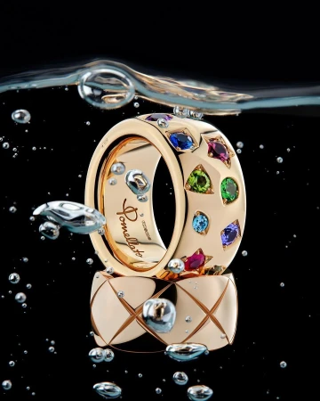 Two gold rings set with colorful gemstones submerged underwater with air bubbles against a black background