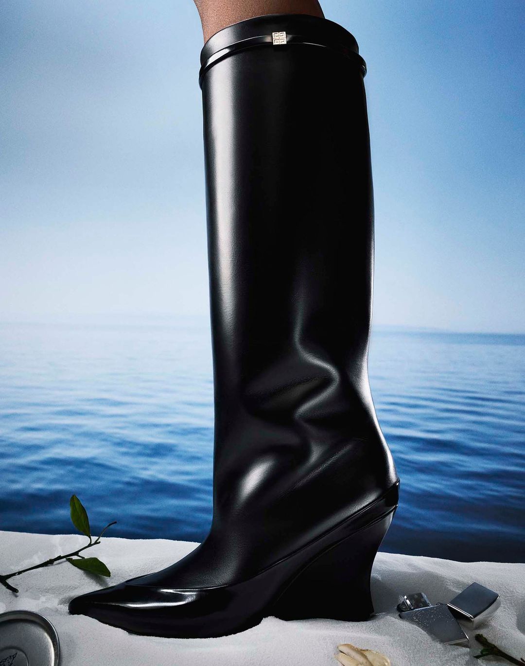 A single black leather knee-high boot with a wedge heel is displayed against a backdrop of water and sky. Fashion footwear concept.