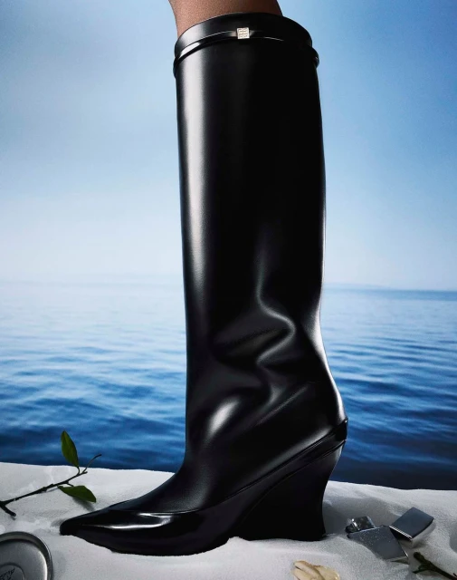 A single black leather knee-high boot with a wedge heel is displayed against a backdrop of water and sky. Fashion footwear concept.