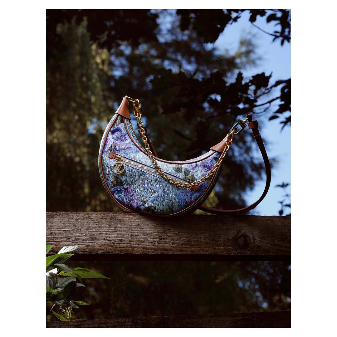 Floral pattern crescent handbag with a gold chain strap sits on a wooden fence with green leaves, set against an out-of-focus tree background.