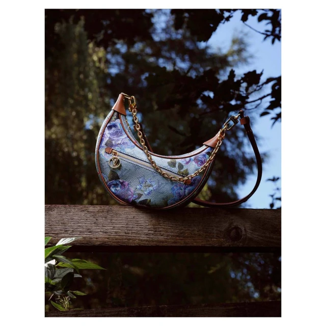 Floral pattern crescent handbag with a gold chain strap sits on a wooden fence with green leaves, set against an out-of-focus tree background.