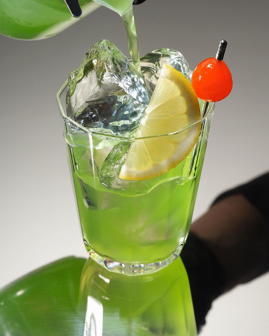 Green liquid is being poured into a faceted glass filled with ice cubes and garnished with a lemon slice and a maraschino cherry.