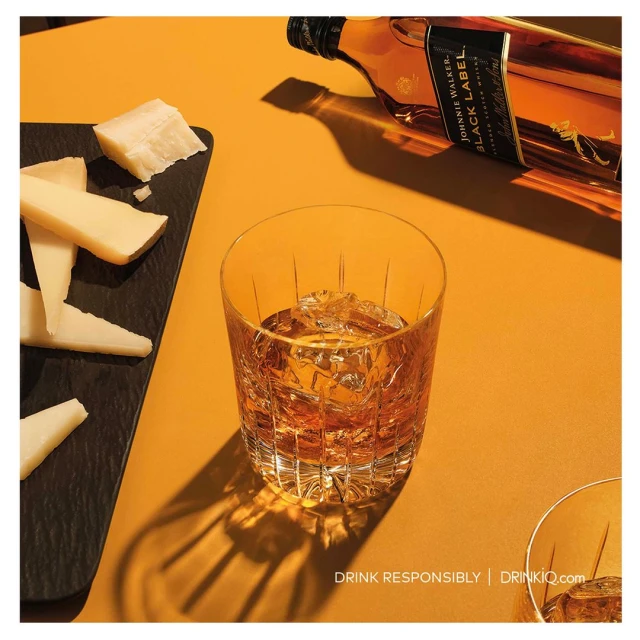 Still life with a glass of whiskey on the rocks, cheese slices on a slate, and a bottle of Johnnie Walker Black Label on a yellow surface.