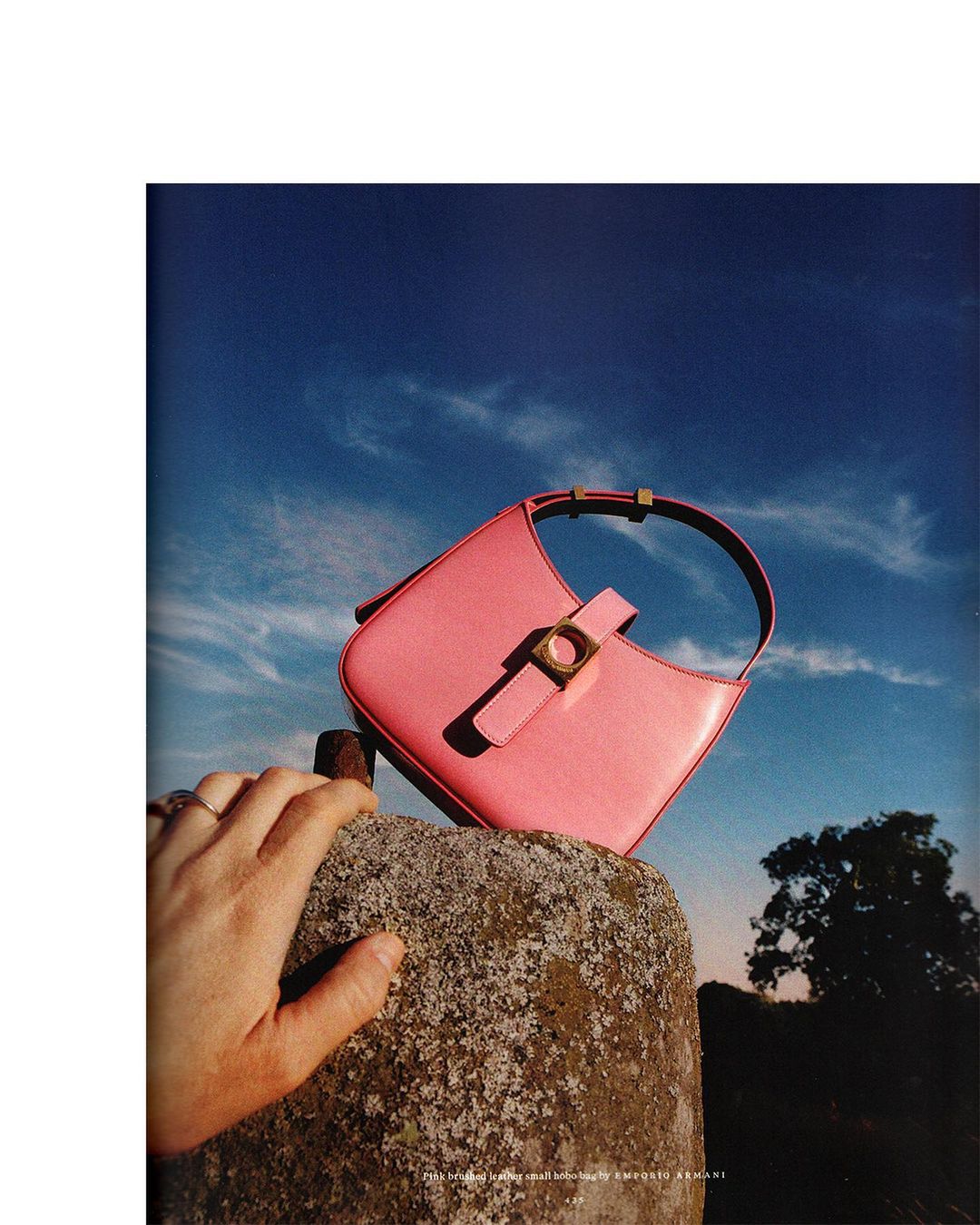 Pink leather handbag with gold hardware displayed outdoors on a stone pillar against a blue sky with a tree in the background.