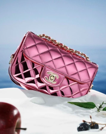 Shiny pink Chanel purse with a gold chain strap on white fabric with a blue background, complemented by an apple and green leaves.