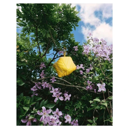 Bright yellow quilted handbag hanging from tree branches among purple clematis flowers against blue sky