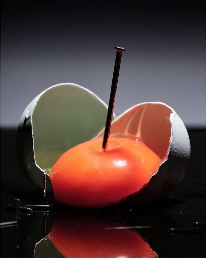 Broken egg with cracked shell and bright orange yolk pierced by nail. Reflective black background and dramatic lighting.