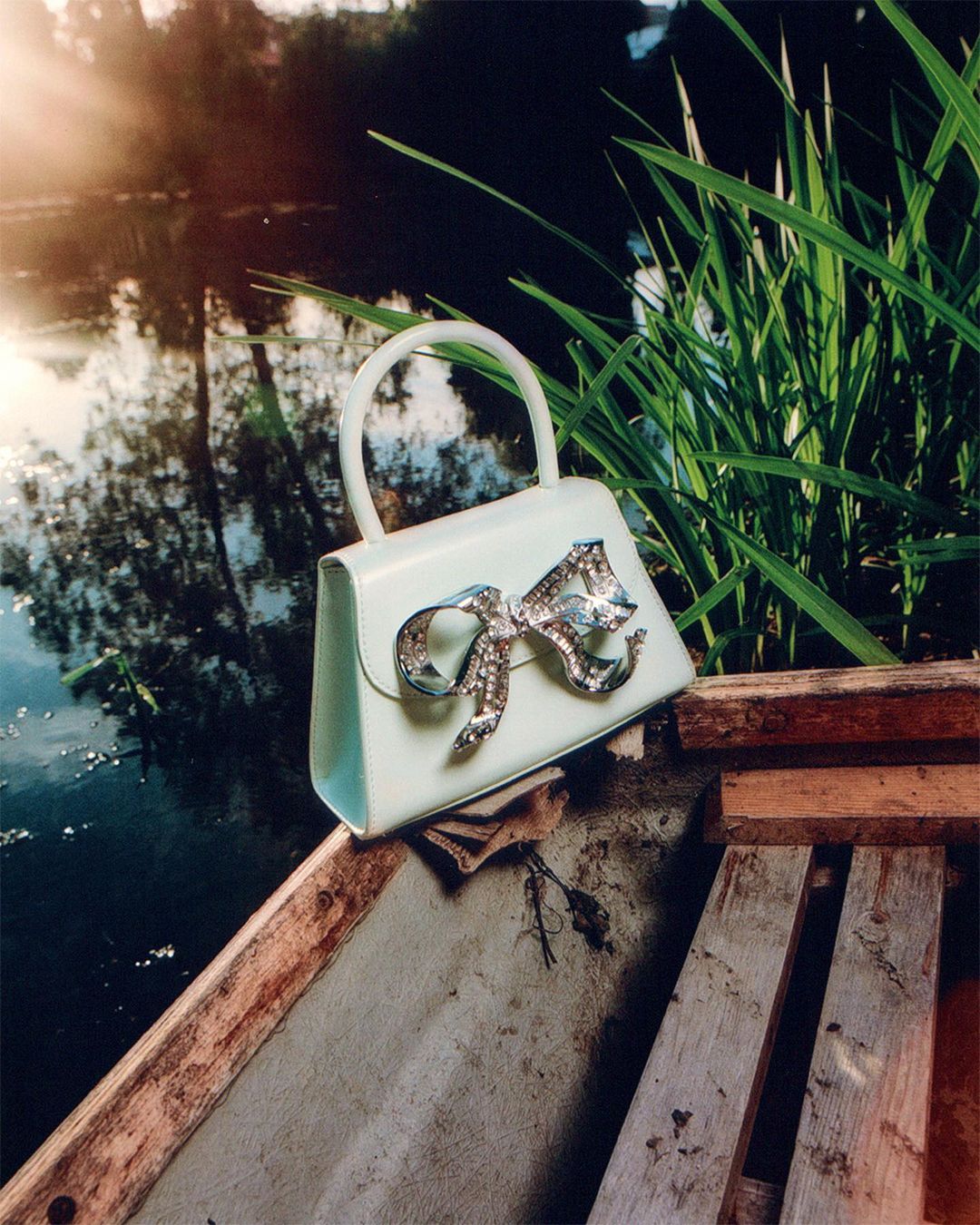 Stylish light-blue handbag with a bejeweled bow resting on a wooden boat, with a serene lake and green reeds in the background.