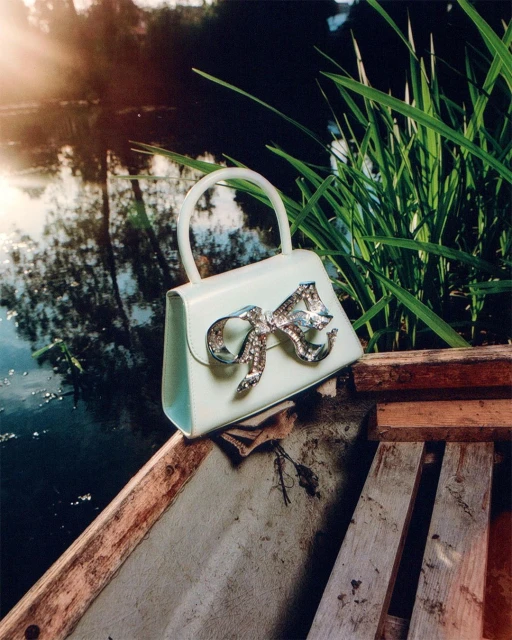 Stylish light-blue handbag with a bejeweled bow resting on a wooden boat, with a serene lake and green reeds in the background.