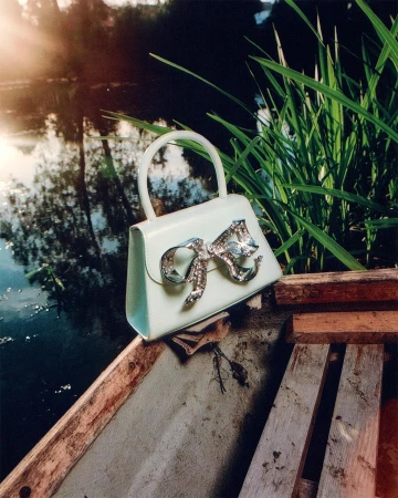 Stylish light-blue handbag with a bejeweled bow resting on a wooden boat, with a serene lake and green reeds in the background.