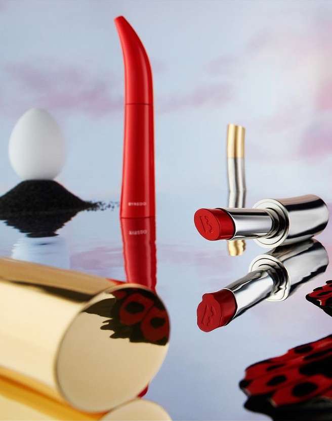 Still life shot of assorted Byredo brand cosmetics including lipstick, mascara, and compact against a mottled studio backdrop.