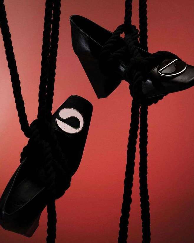 Two black leather block heel shoes are suspended with thick black rope against a muted reddish-brown backdrop in a studio setting.