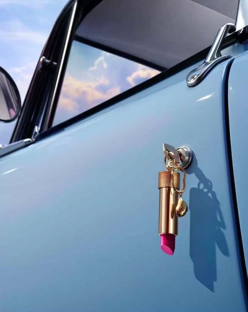 Lipstick keychain hanging from a light blue car door with clouds reflecting in the window, creating a blend of beauty and automotive themes.