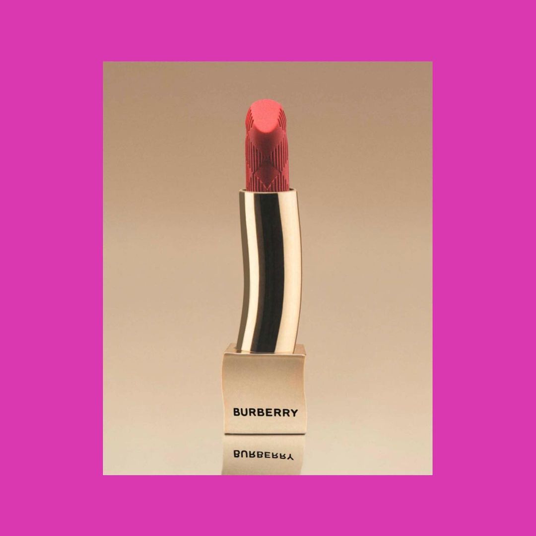 Close-up studio shot of a Burberry lipstick tube with the brand name visible. The lipstick is a coral color and gold.