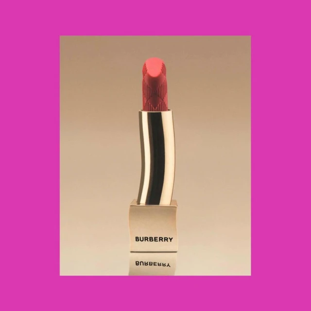 Close-up studio shot of a Burberry lipstick tube with the brand name visible. The lipstick is a coral color and gold.