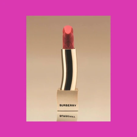 Close-up studio shot of a Burberry lipstick tube with the brand name visible. The lipstick is a coral color and gold.