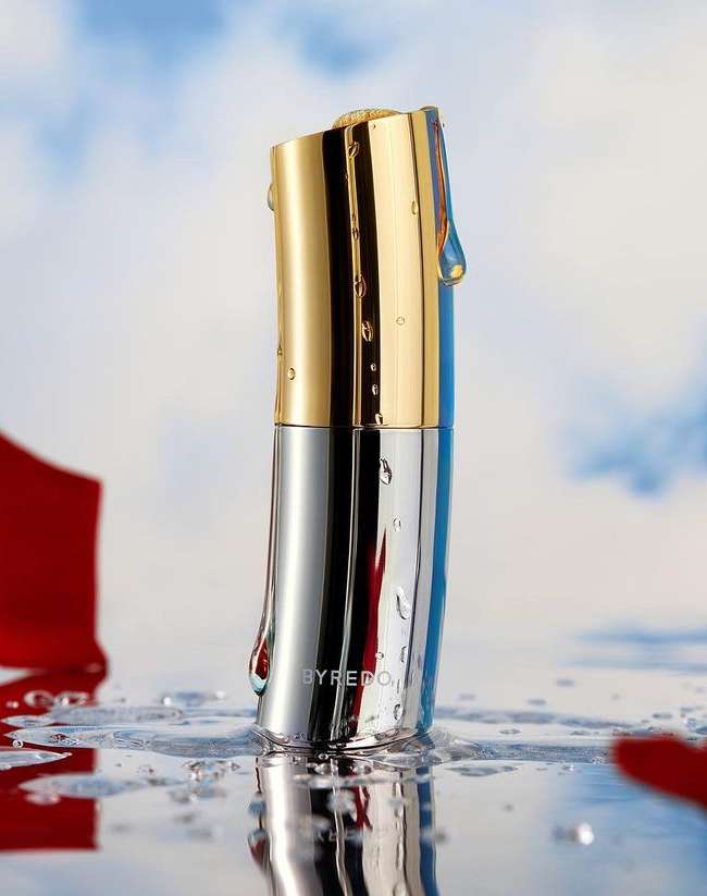 Studio shot of gold and silver Byredo fragrance bottle sitting in shallow water against blurred sky and clouds backdrop.
