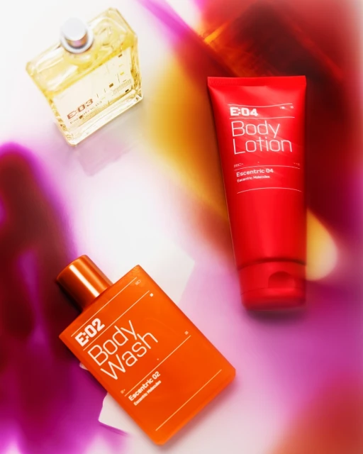 Still life of E-04 Body Lotion in red tube, E-02 Body Wash in orange bottle, and E-03 fragrance in clear bottle against colorful background.