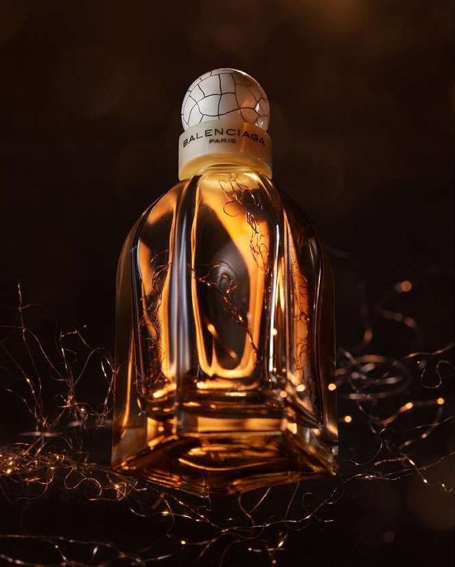 Elegant glass Balenciaga perfume bottle with decorative cap and accents, reflecting golden light in a dark, moody fragrance ad.