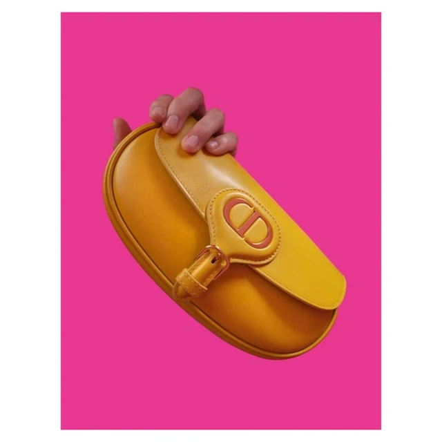 Close-up studio shot of a yellow leather handbag with a logo detail, held by a hand, set against a vibrant pink background.