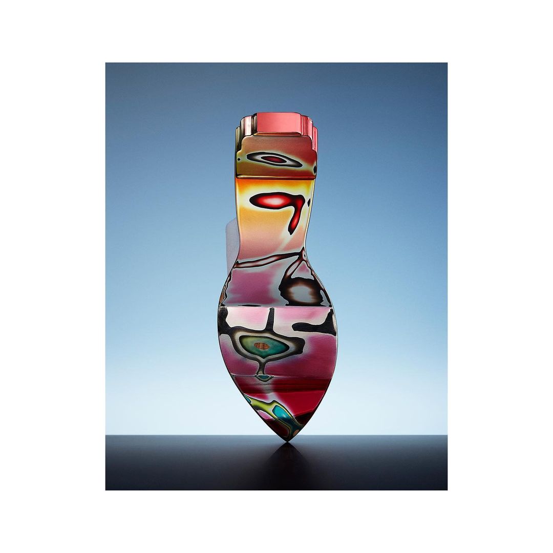 Abstract art glass vase in gradient hues of pink, red, and yellow with abstract patterns stands on a black surface against a blue background.
