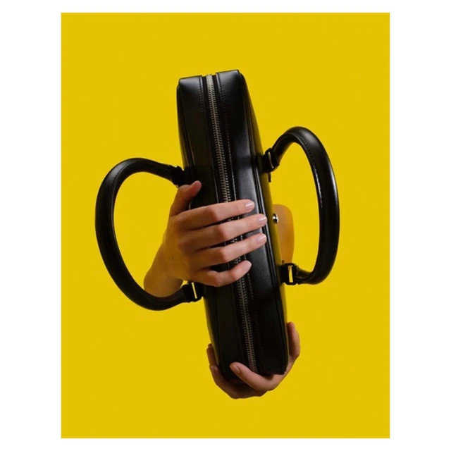 Close-up of a black leather handbag held by a person with manicured nails against a vibrant yellow background, fashion accessory.