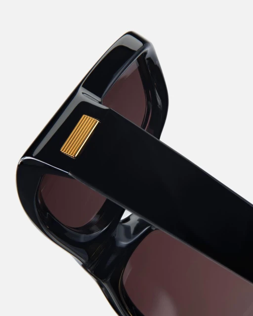 Angled view of black, square-frame sunglasses with a gold accent on the temple, showcasing the lens tint and design against a white background.