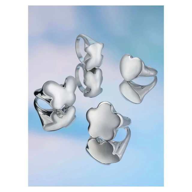 Close-up of silver rings with heart, flower, and bear designs displayed on a reflective surface with a gradient blue background.