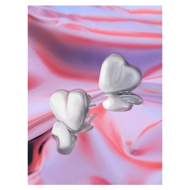 Two heart-shaped silver stud earrings stand on a reflective surface with a swirled pink, purple, and white background, in a studio shot.