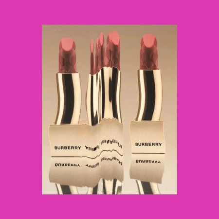 Three Burberry lipsticks are displayed, with a distorted reflection of one, set against a light gradient background and a pink square.