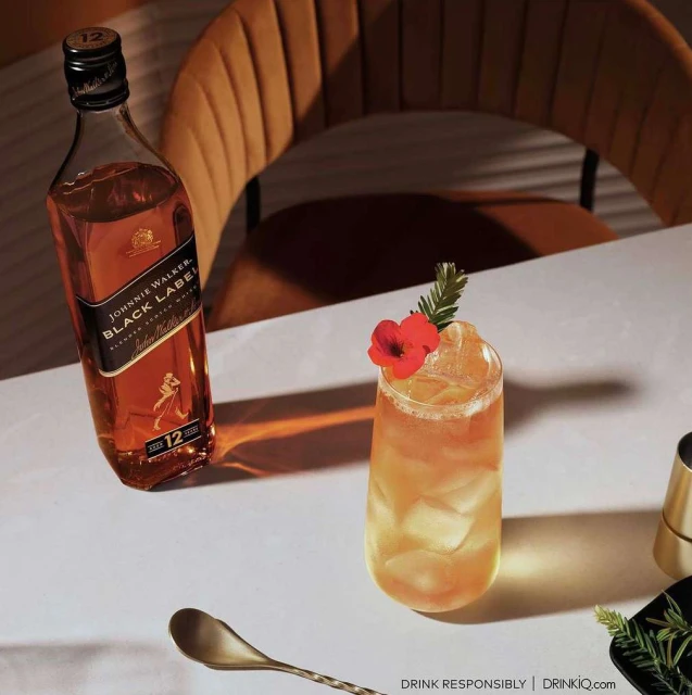 Whiskey drink still life: Johnnie Walker Black Label bottle and cocktail with red flower garnish, sitting on a white table indoors.