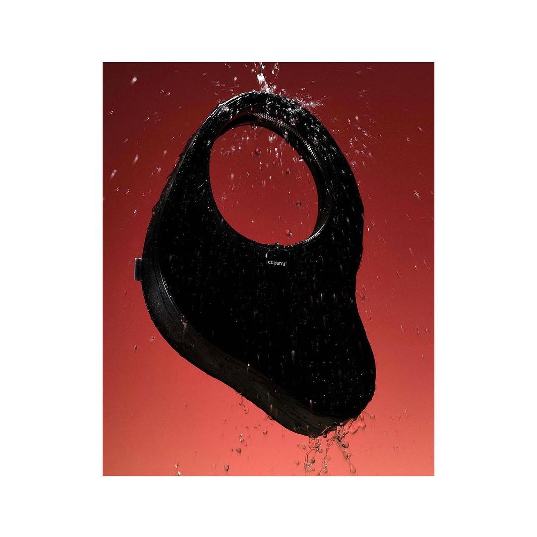 Black handbag with a circular handle stands out against a rich red background, enhanced by water droplets adding a dynamic visual texture.