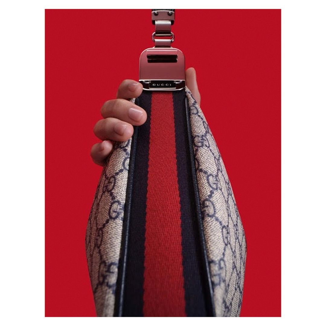 Close-up of a Gucci bag being held, featuring a beige pattern, red and black strap, and a metal buckle against a vibrant red background.