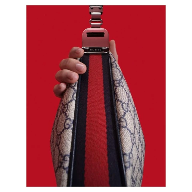 Close-up of a Gucci bag being held, featuring a beige pattern, red and black strap, and a metal buckle against a vibrant red background.
