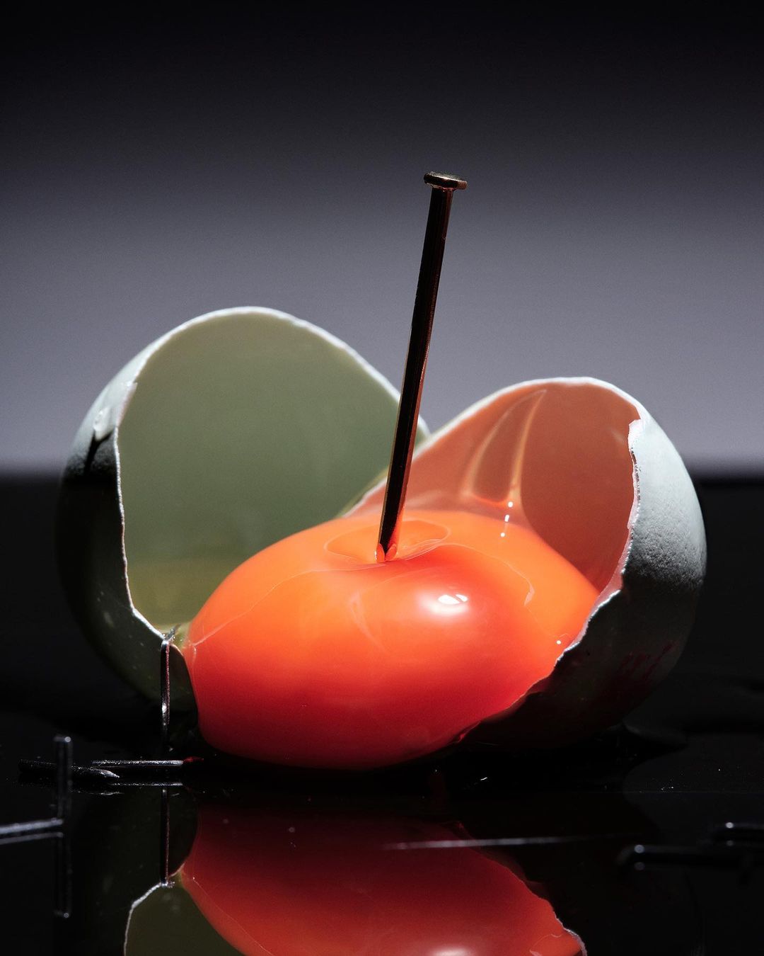 Cracked egg with dripping yolk pierced by a nail, against a dark background with reflections. Food photography concept.