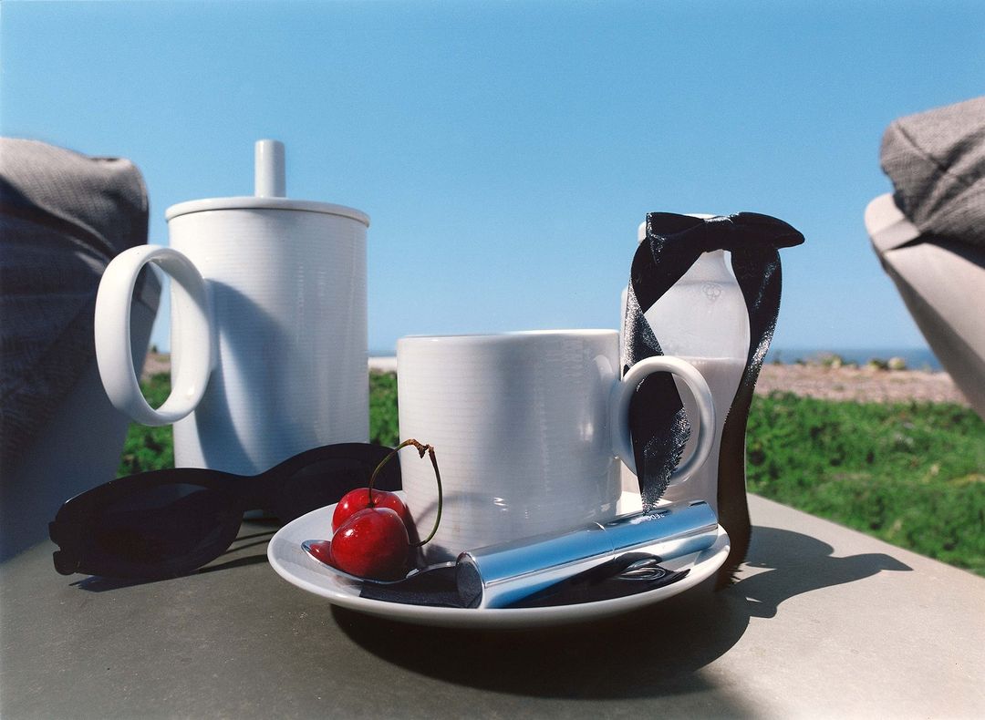 Outdoor still life features white cups, cherries, sunglasses, and milk against blue sky and green foliage background on a sunny day.