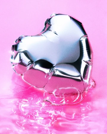 Silver heart-shaped balloon floating on a pink reflective surface. The balloon reflects the surrounding light and colors with a metallic sheen.