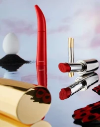 Red and silver lipstick tubes and curved mascara on a reflective surface with an egg and black sand under a pastel sky.