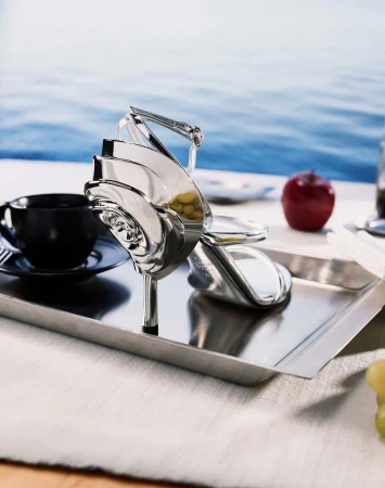 Silver metallic heeled sandal shaped like a rose on a metal tray beside a black teacup and red apple with water in background