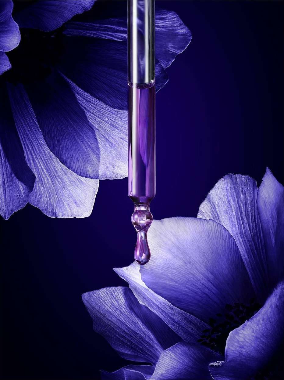 Close-up studio shot of a silver and clear dropper with purple liquid, with a drop forming, set against a floral and purple background.