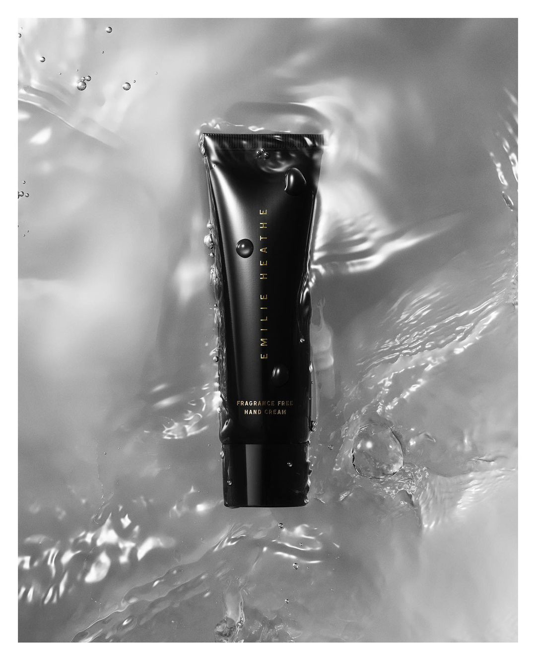 Black tube of Emilie Heathe Fragrance Free Hand Cream surrounded by water droplets on a light grey background, product shot.