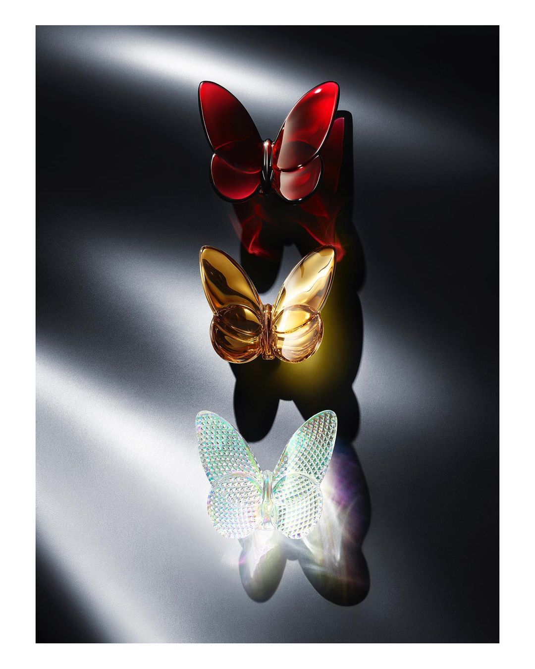 Three crystal butterfly figurines in red, gold and white are vertically stacked on a glossy surface against a dark gray background.